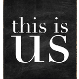 A black and white wood block sign with the text 'This Is Us' in the center. Available at Be Charmed