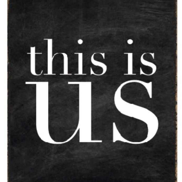 A black and white wood block sign with the text 'This Is Us' in the center. Available at Be Charmed