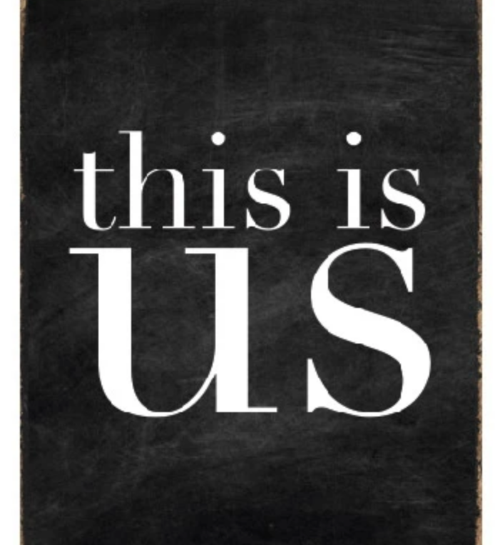 A black and white wood block sign with the text 'This Is Us' in the center. Available at Be Charmed
