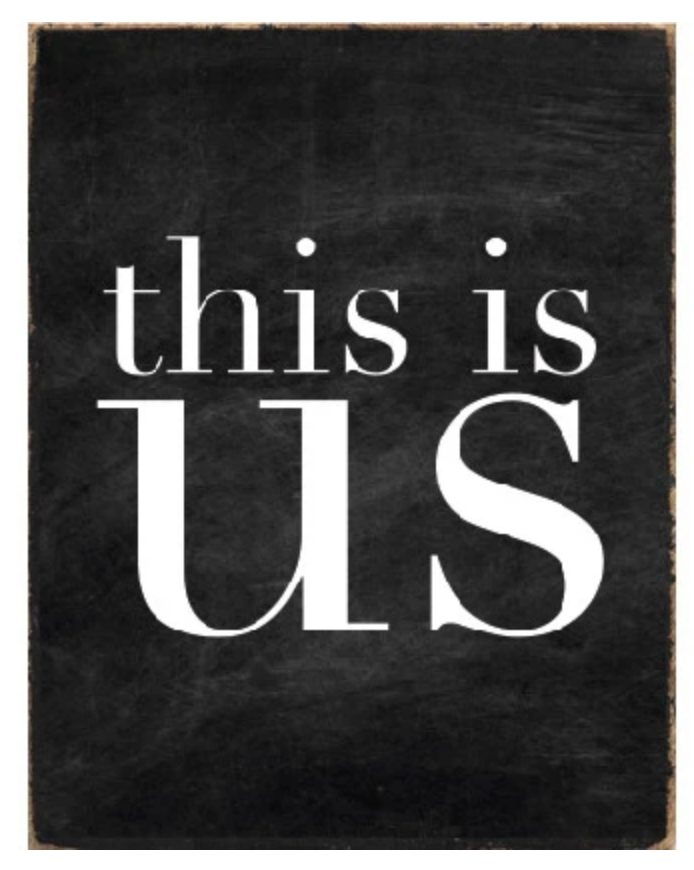 A black and white wood block sign with the text 'This Is Us' in the center. Available at Be Charmed