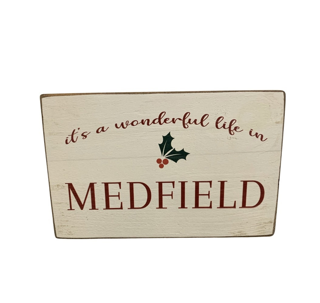 A beige rustic wooden block with the text 'It's a wonderful life in Medfield' and Christmas decorations. Available at Be Charmed