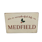 A beige rustic wooden block with the text 'It's a wonderful life in Medfield' and Christmas decorations. Available at Be Charmed