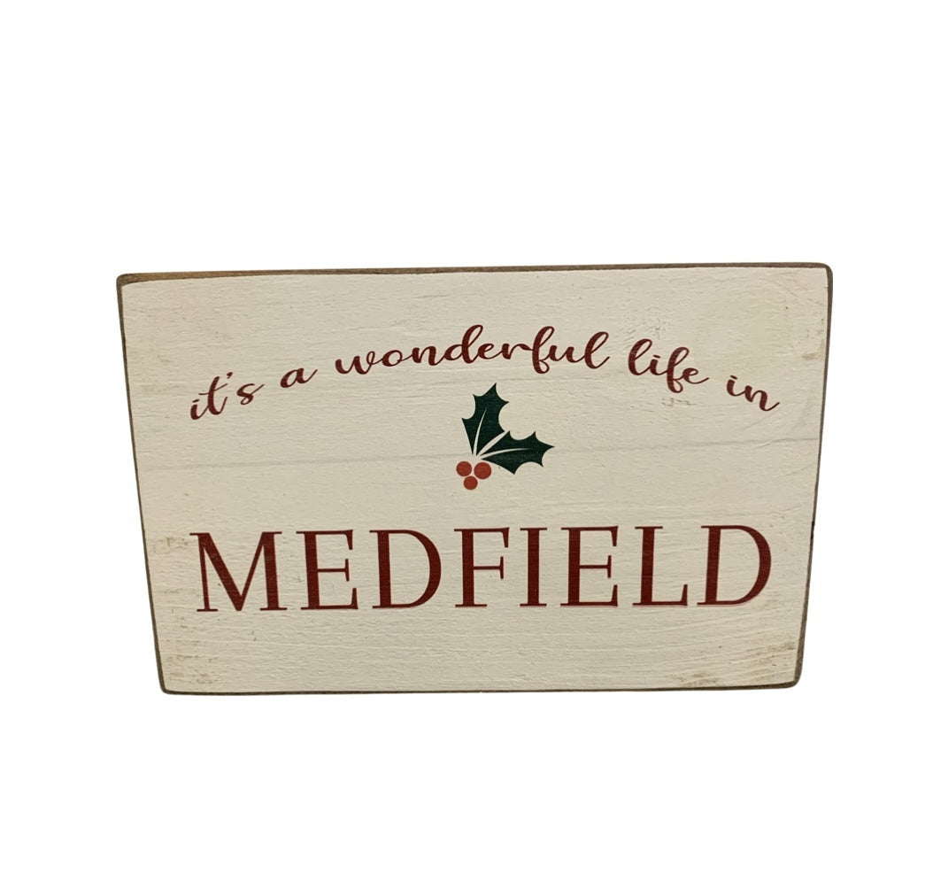 A beige rustic wooden block with the text 'It's a wonderful life in Medfield' and Christmas decorations. Available at Be Charmed