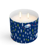 A clear Scout candle with a blue label featuring a Christmas cabin and tree pattern, with a lit wick. Available at Be Charmed
