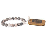 A grey stone stacking bracelet with a label tagged 'stone of plentitude'. Available at Be Charmed