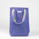 A purple woven Scout tote bag with a multicolored striped handle. Available at Be Charmed