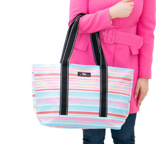 A woman holding a multi striped Scout tote bag with black straps and a front brand label. Available at Be Charmed