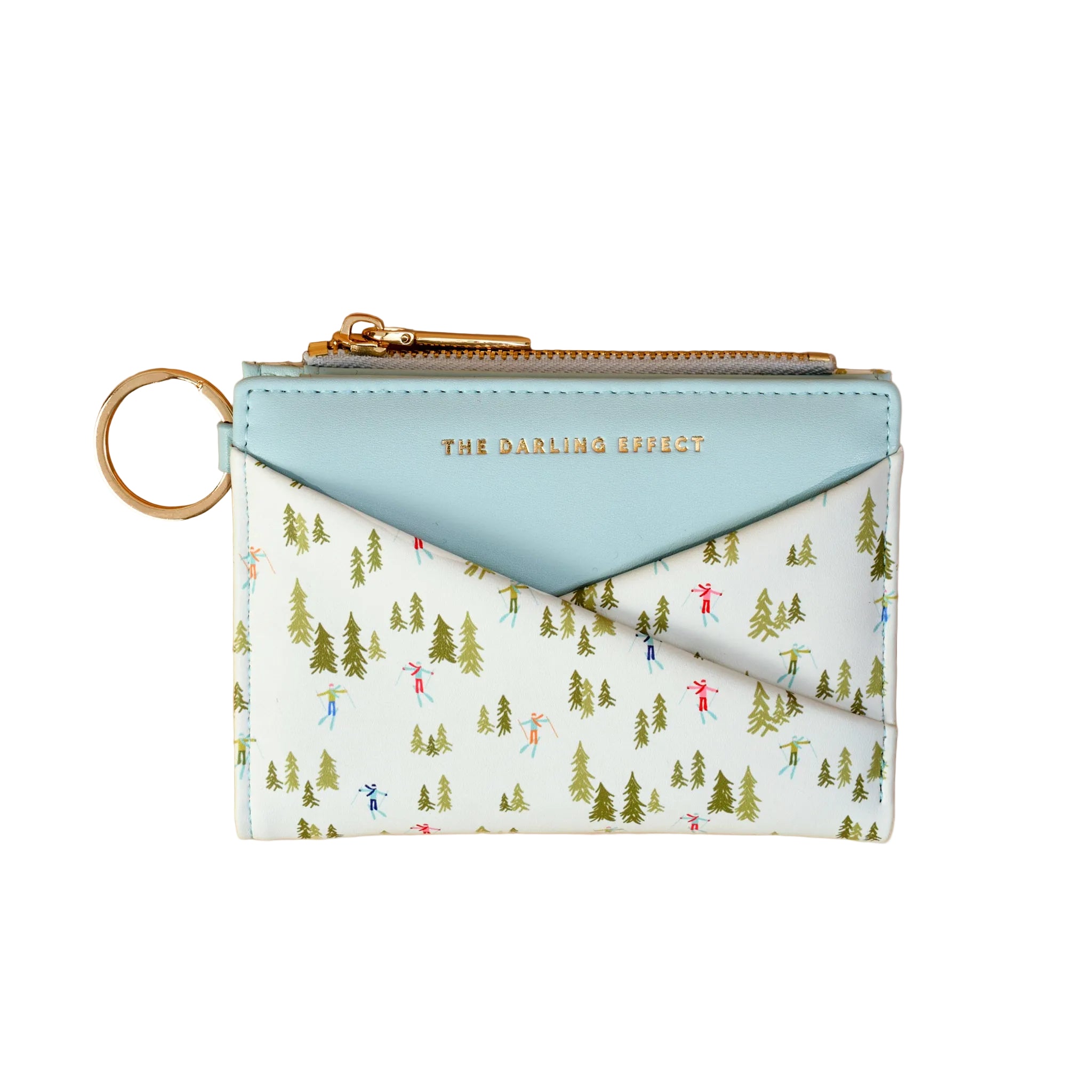 A zip-around card holder wallet by The Darling Effect, featuring a light blue leather flap with gold lettering and a white body printed with a green pine tree and colorful skier pattern, complete with a gold key ring attachment. Available at Be Charmed