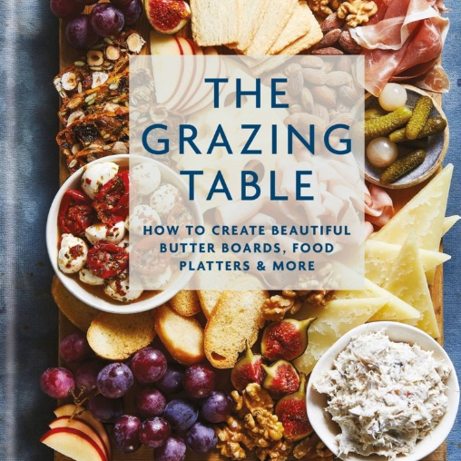 Book cover of 'The Grazing Table' with a wooden board filled with food items on a blue background. Available at Be Charmed