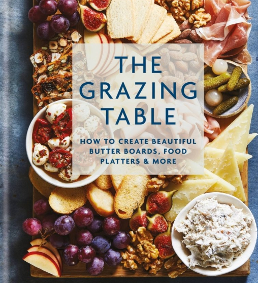 Book cover of 'The Grazing Table' with a wooden board filled with food items on a blue background. Available at Be Charmed