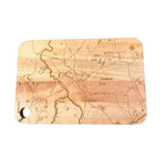 A Well Told rectangular wooden cutting board with rounded corners, featuring an engraved topographic map design in light and dark brown tones, and a hanging hole on the bottom left, shown against a plain white background. Available at Be Charmed