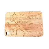 A Well Told rectangular wooden cutting board with rounded corners, featuring an engraved topographic map design in light and dark brown tones, and a hanging hole on the bottom left, shown against a plain white background. Available at Be Charmed